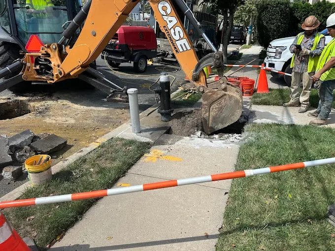 Sidewalk construction scene with excavator digging, surrounded by workers and safety cones. Urban infrastructure maintenance site.