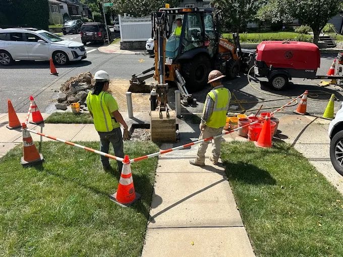 Construction workers using excavator for sidewalk repair, safety cones, and barriers in residential street, August 2024.