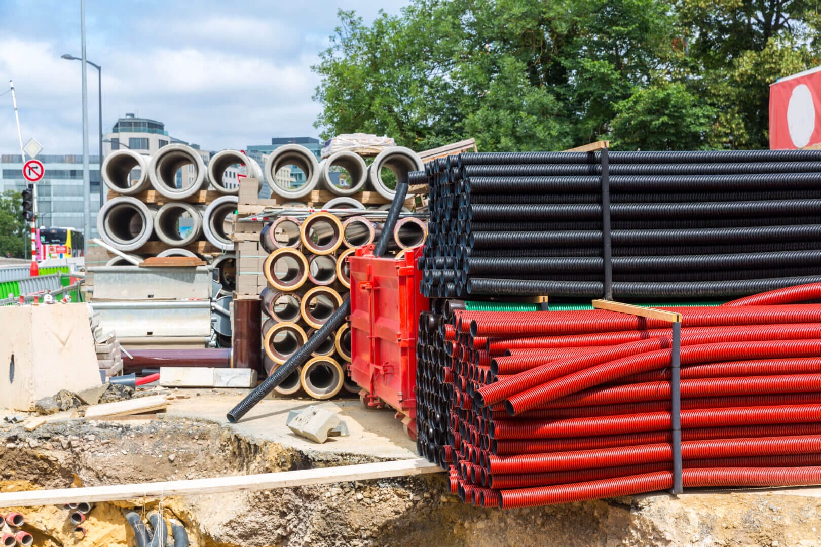 Construction site in Europe with stacks of building materials, including red and black pipes, concrete sections, and urban background.
