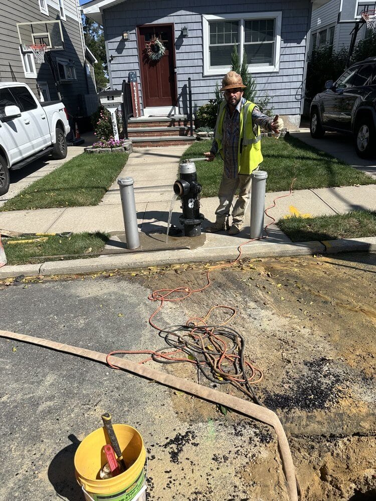Construction worker repairing fire hydrant on residential street, wearing safety vest and hat, tools and hoses visible.