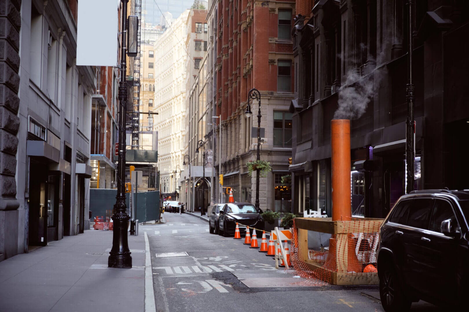 Steaming ventilation pipe on narrow New York City street with parked cars, urban metropolis atmosphere.
