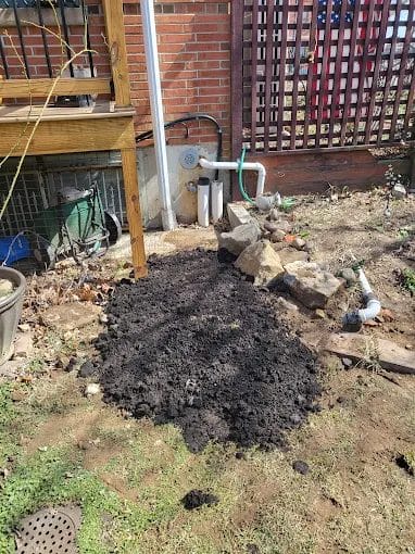 Garden soil preparation near brick wall and wooden deck with scattered rocks and plumbing pipes in backyard.