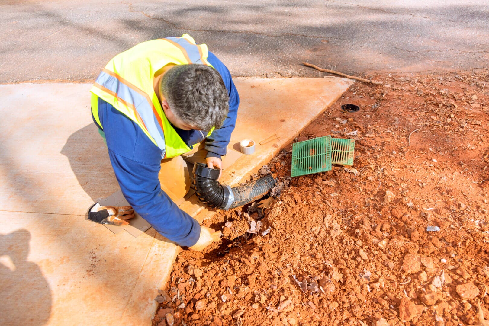 Worker repairing drainage system at residential construction site with tools and safety vest.